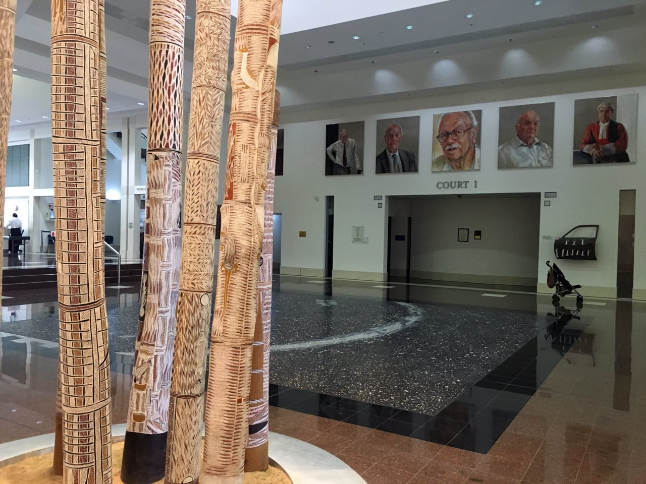 Image 02: View of the central hall at the Supreme Court in Darwin. Visible are portraits by Danielle Bergstrom, poles from a Wukidi ceremony in memory of the Yolŋu man Dhäkiyarr and a Utopia Car Door by Elsie Kemarre. Photo credit: Alana Hunt
