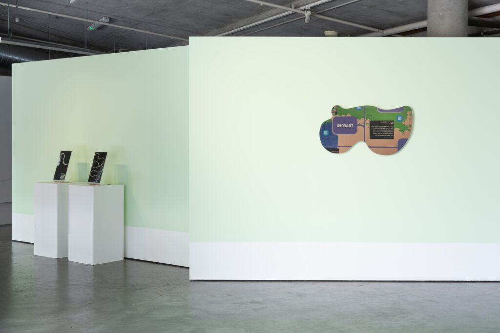 Installation view of the exhibition. Against a mint green wall with white trim are, on the left, two plinths with two vertical rectangle mirrors with etching. On the right hand side is a mirror, shaped in a blob-like, amorphous way. The mirror is reflecting the vinyl wall work.