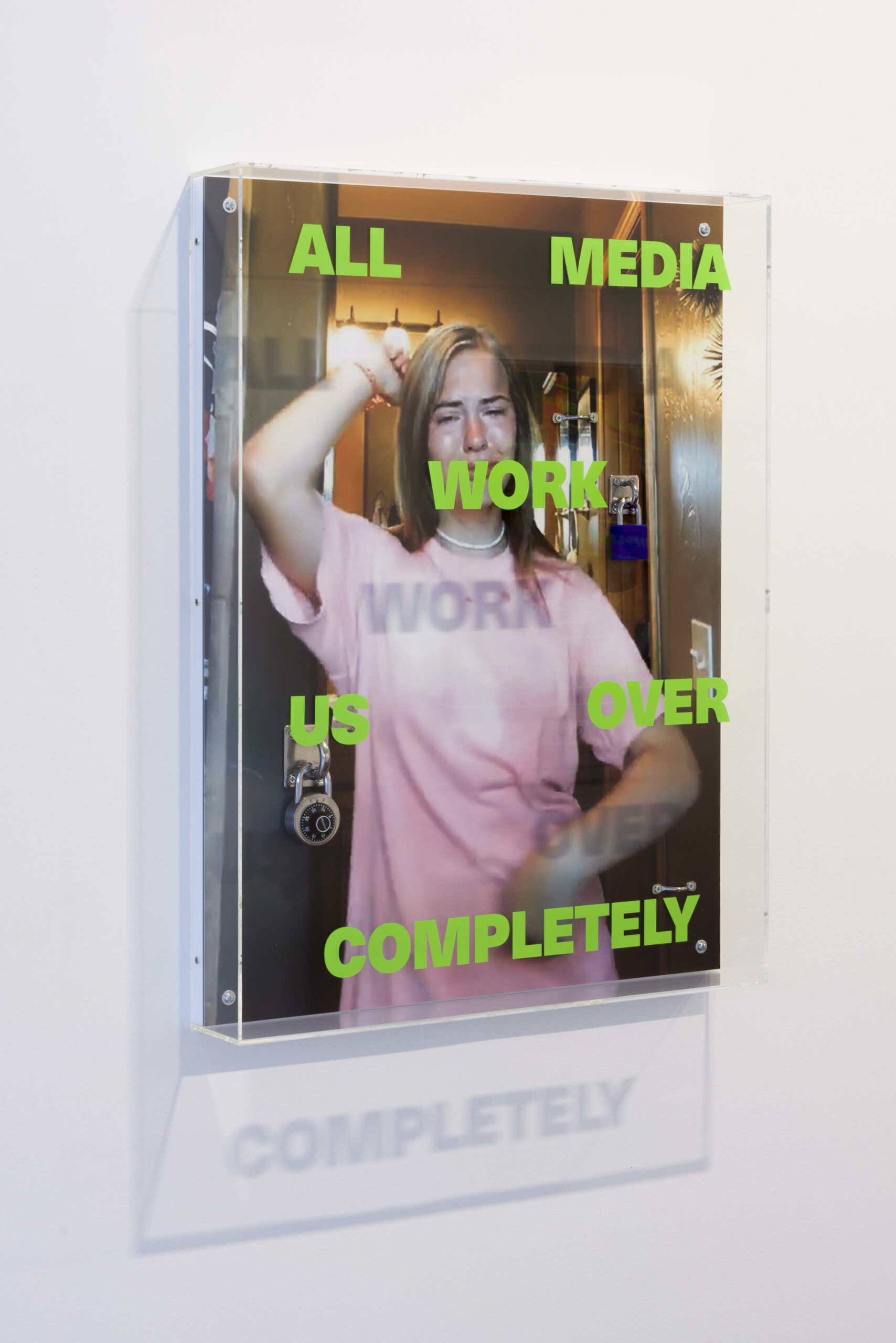Documentation of an artwork by Dan Bourke. Portrait oriented, the photo print is incased in an acrylic frame. The image is a screen shot of a woman with blonde hair wearing a pink t-shirt dancing for a TikTok video while crying. Green vinyl text in all capitals is laid on the frame reading "ALL MEDIA WORK US OVER COMPLETELY"